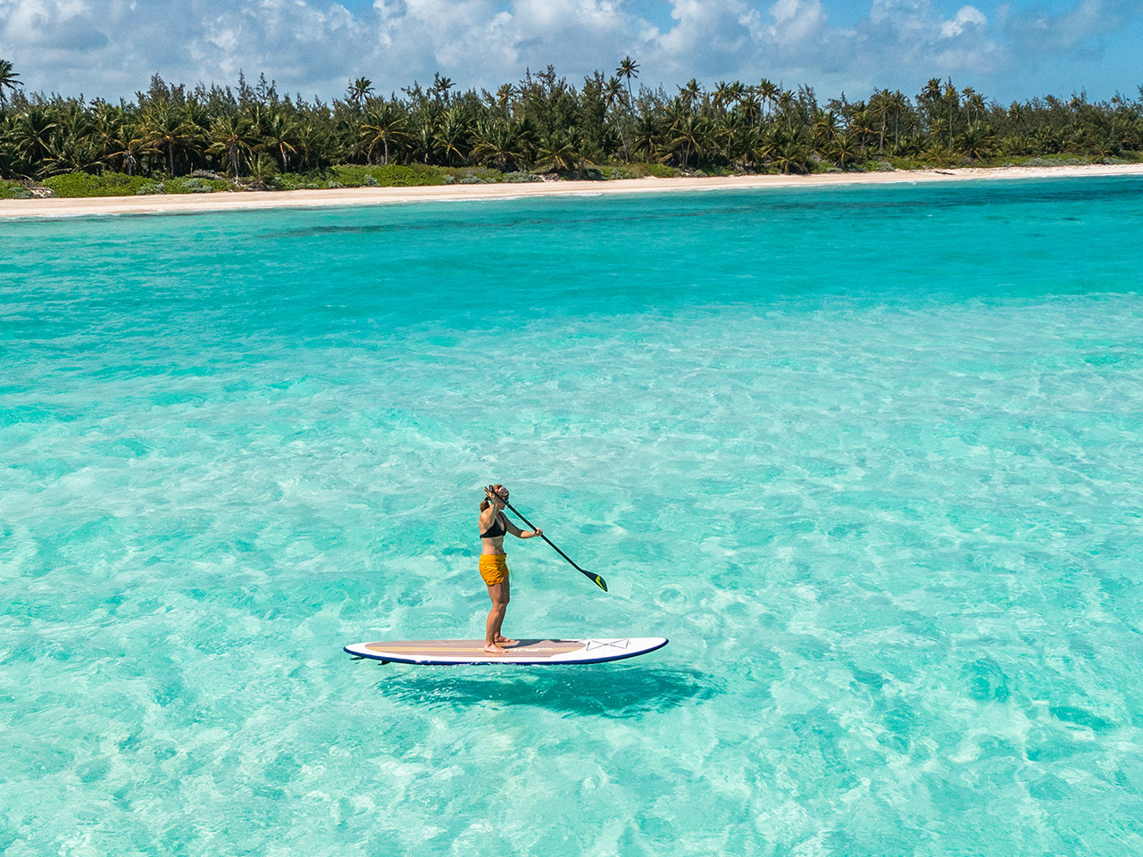 A person paddleboards on clear turquoise water near the beach at Caerula Mar Club, a luxury resort on South Andros Island, Bahamas.