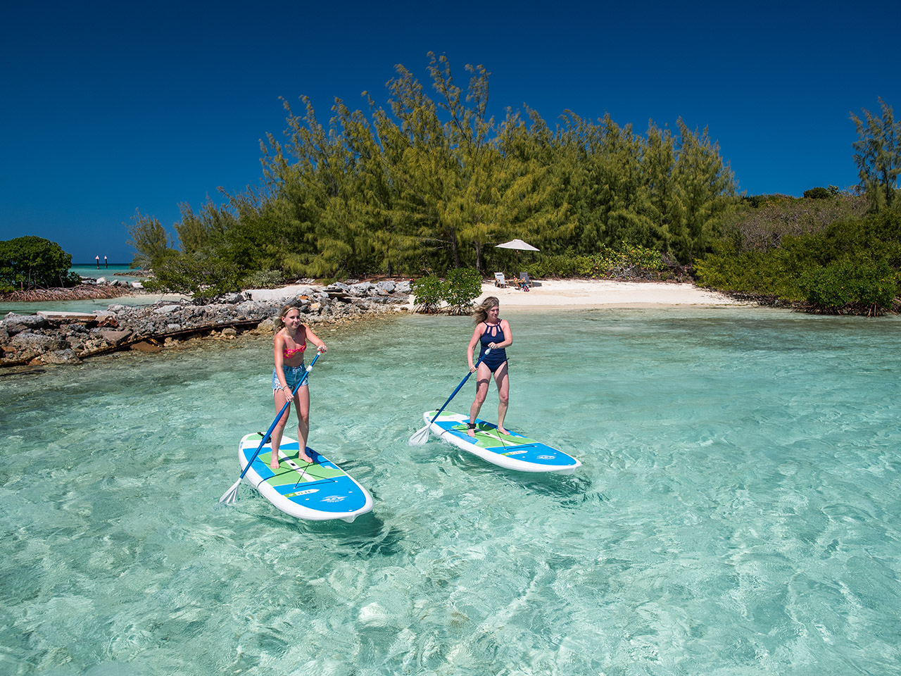 Two women paddleboard on turquoise water near the sandy, lush shore at Caerula Mar Club, luxury resort in South Andros, Bahamas.