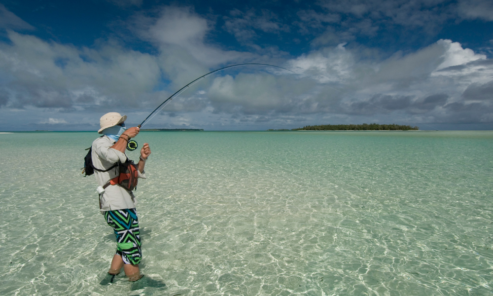 A guest in colorful shorts and a hat stands knee-deep in the pristine, shallow waters at Caerula Mar Club on South Andros Island, fly fishing under a partly cloudy sky with a lush green island visible on the horizon.