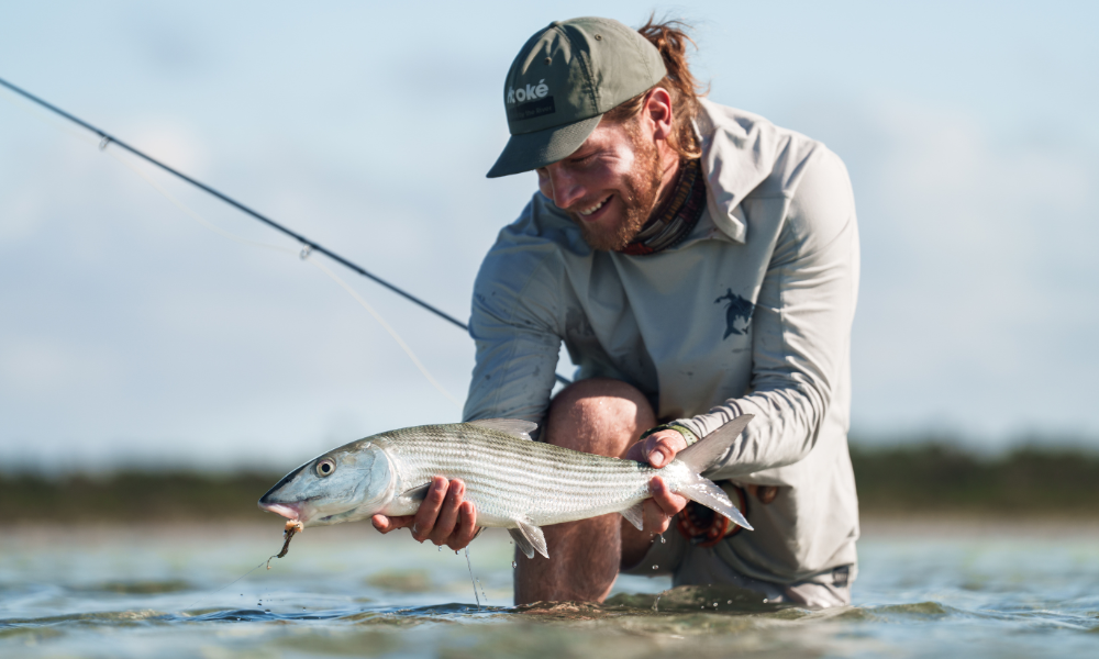 Guest enjoys fishing in the clear, shallow waters at Caerula Mar Club on South Andros Island, smiling while holding a silver, striped fish with a fishing rod visible in the background.